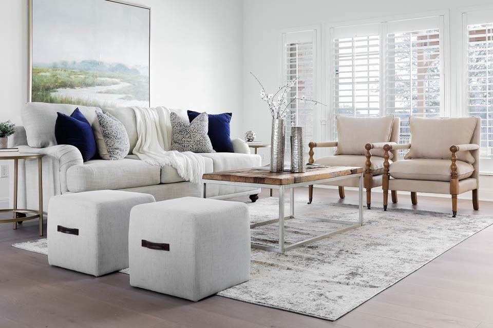 wood flooring with patterned area rug in living room with grey couch, two beige chairs, and two grey fabric ottomans with metal coffee table.